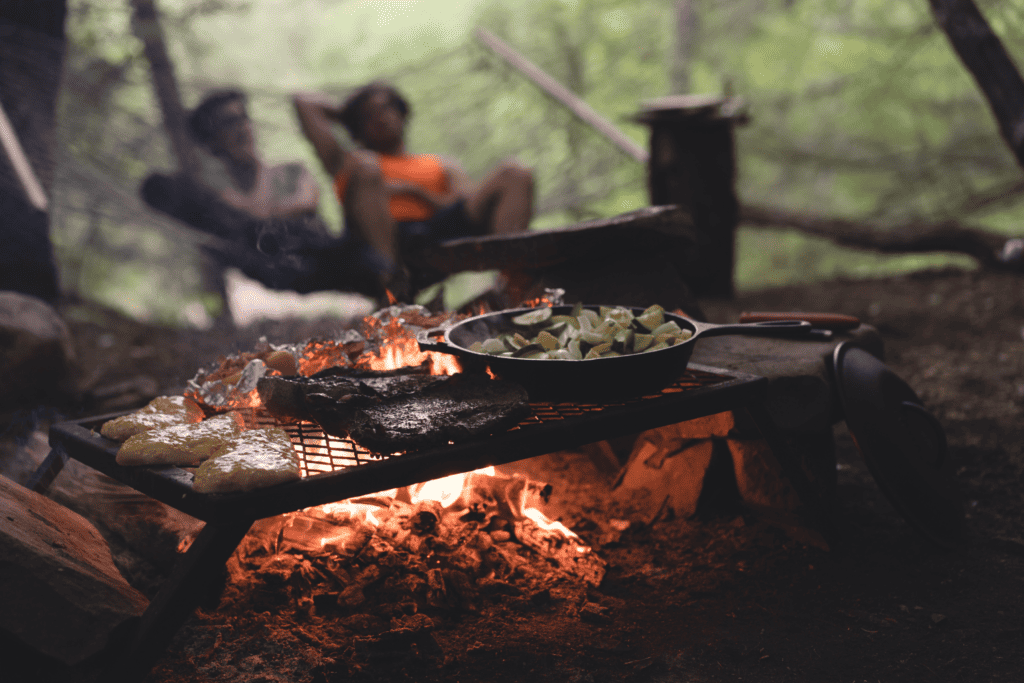 Food cooking on an outside fire with two people relaxing in the background. Photo by Myles Tan on Unsplash