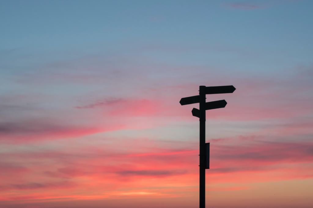 A signpost pointing in different directions with the sunset behind it. Photo by Javier Allegue Barros on Unsplash
