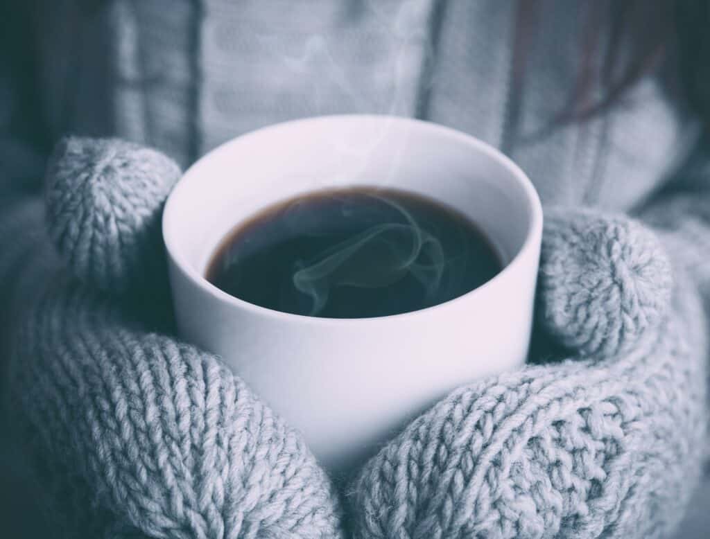 Someone holding a hot cup of tea/coffee while wearing a pair of mittens. Photo by Alex Padurariu on Unsplash