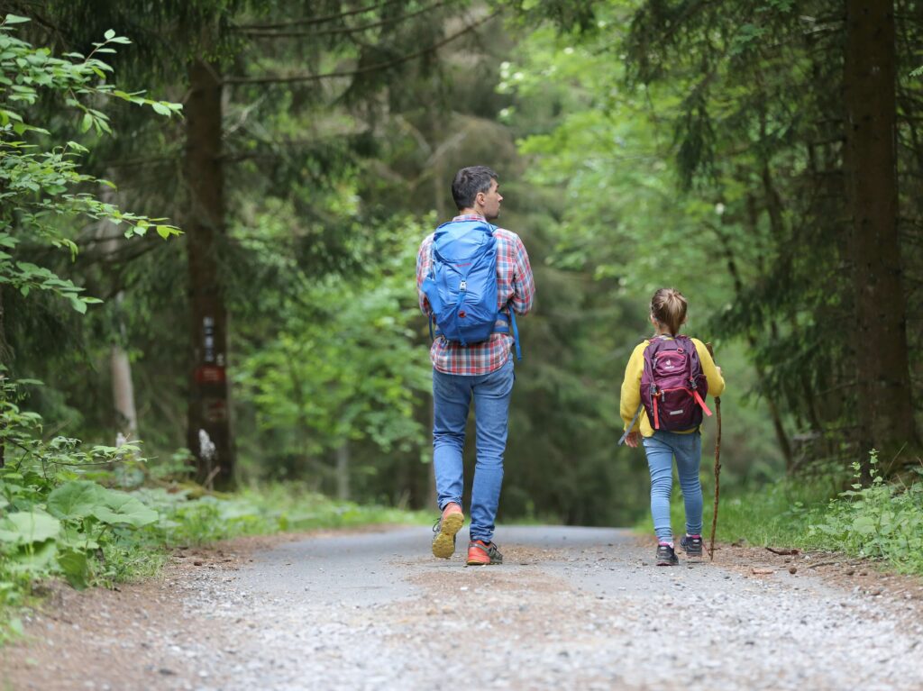 A father and daughter hiking through a forest, both of them wearing rucksacks. Photo by Juliane Liebermann on Unsplash