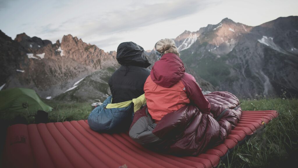 Two people sitting outside in their sleeping bags on top of sleeping mats, overlooking a mountain side. Photo by Lucas Canino on Unsplash
