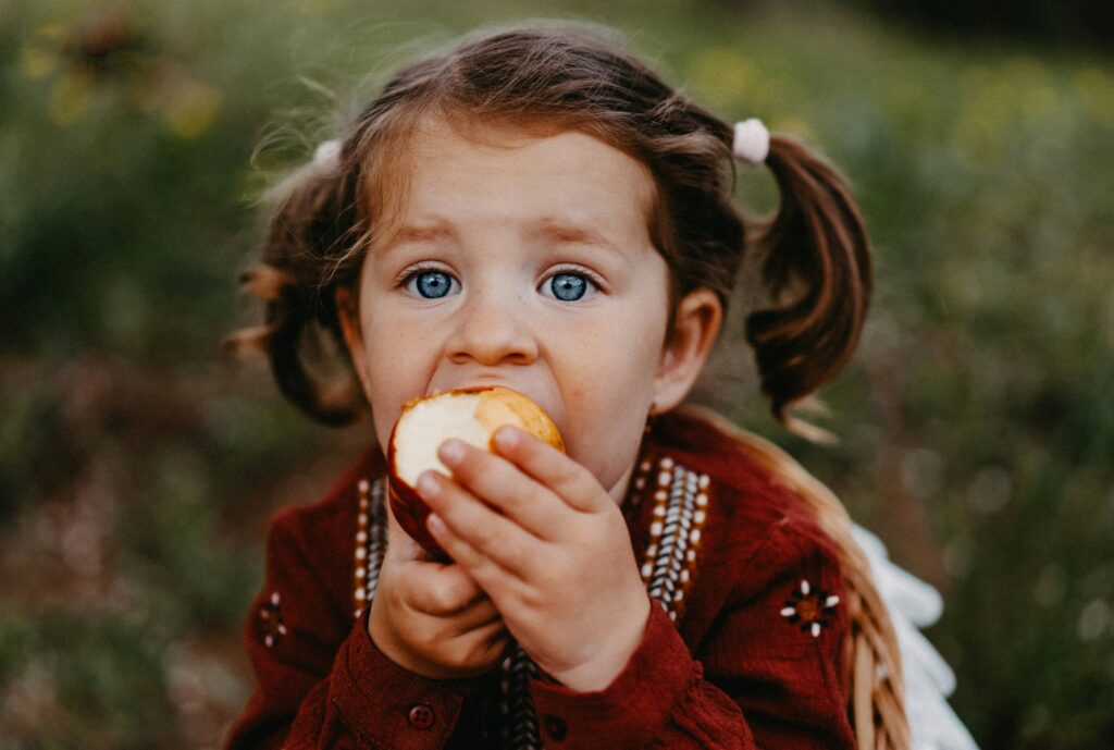 A young girl with blue eyes and brown hair eating an apple. Photo by Raymond Petrik on Unsplash