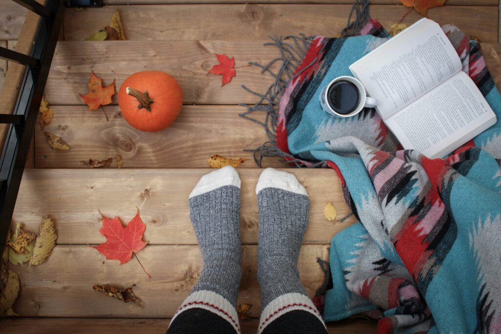 Someone wearing a pair of warm socks with a book, cup of tea/coffee, a blanket, and a small orange pumpkin on wooden steps. Photo by alex geerts on Unsplash
