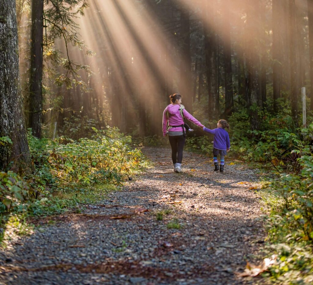A mother and child walking hand-in-hand along a forest path, looking at each other. Photo by James Wheeler on Unsplash