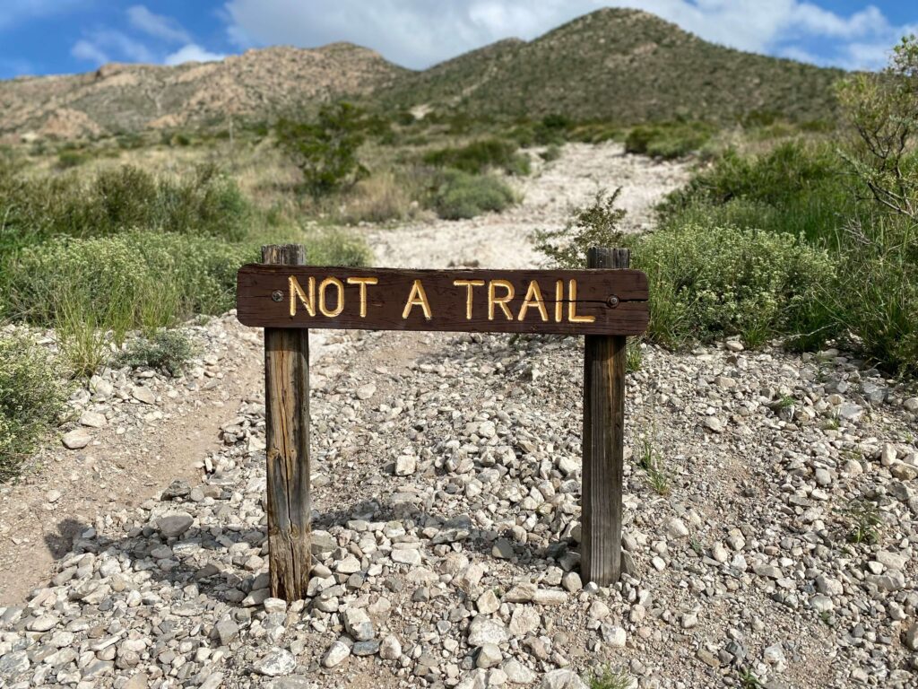 "Not a Trail" sign in the foreground along a gravel path with mountains in the background. Photo by Ma lie on Unsplash