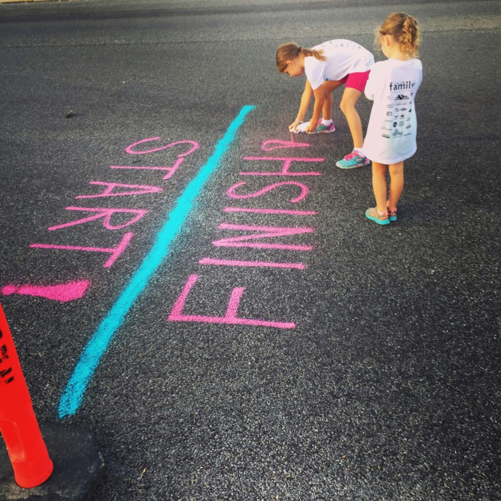 Two young girls spraying "Start" and "Finish" on the tarmac with a blue line between the two words. Photo by Adam Winger on Unsplash