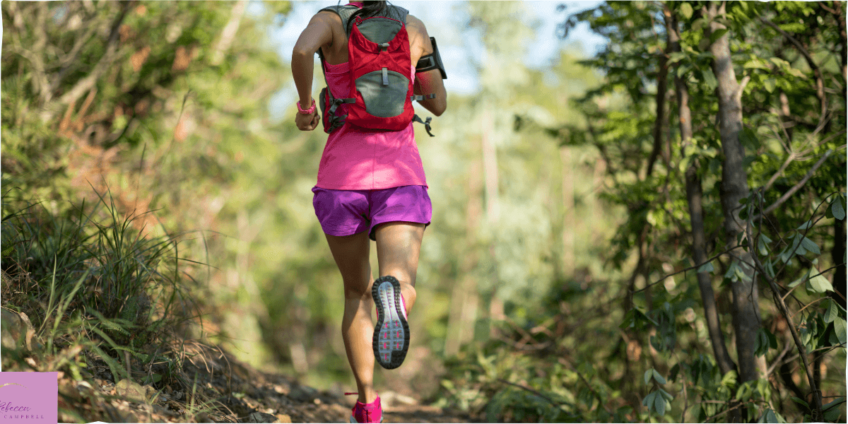 A trail runner in pink and purple running along a forested path. Source: Canva