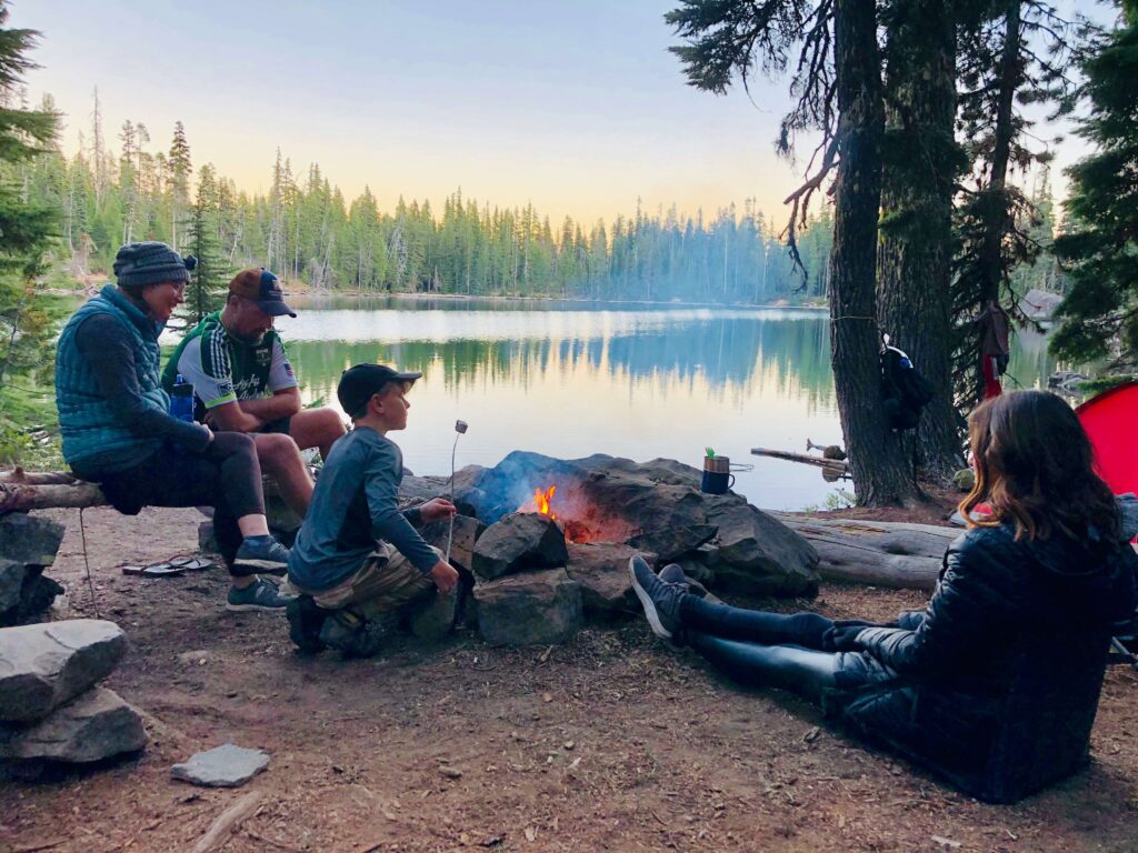 A family camping near a lake around a campfire, eating marshmallows. Photo by Brooks Rice on Unsplash
