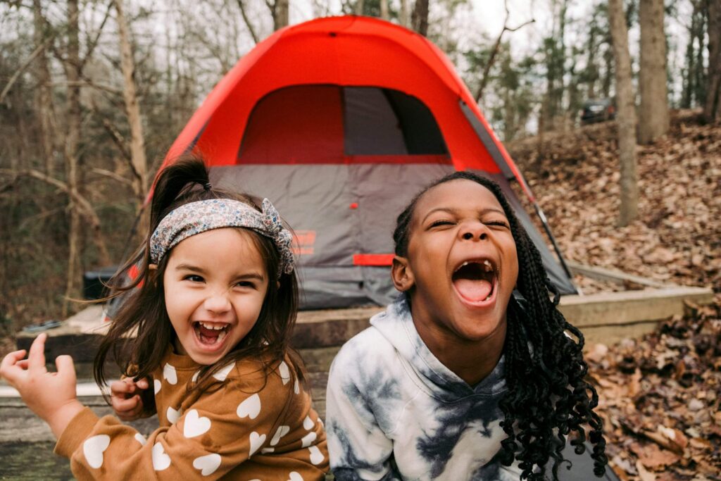 Two girls laughing while sitting outside in a forested area with a red tent behind them. Photo by Colin + Meg on Unsplash