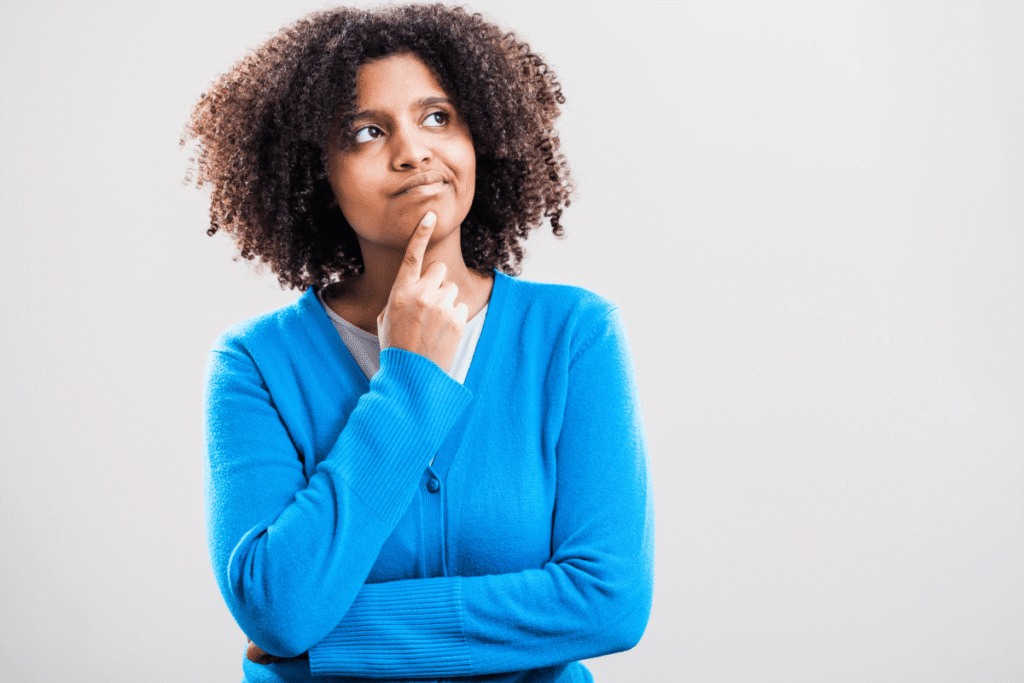 A woman in a blue cardigan with one arm crossed and the other hand resting under her chin, with a thinking face on. Source: Canva